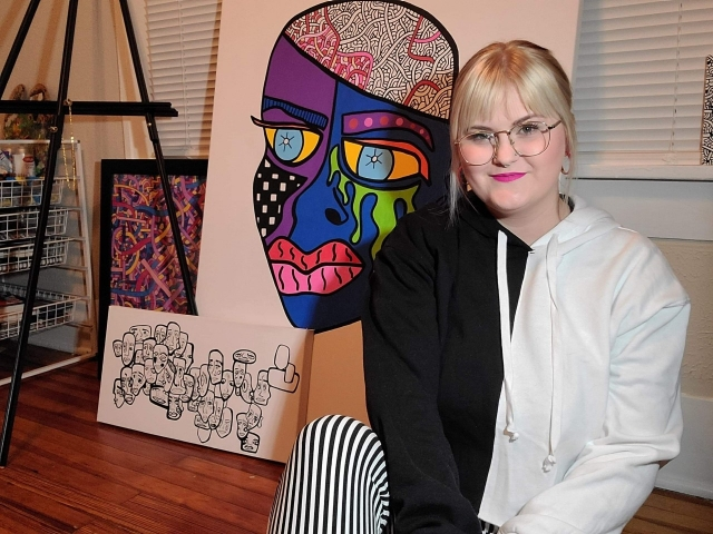 A blonde woman sitting in her studio. She is wearing a half white and half black top. She sits in front of her paintings.