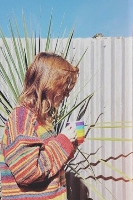 Side view of a person with orange hair, a striped colorful sweater holding a rainbow mug.