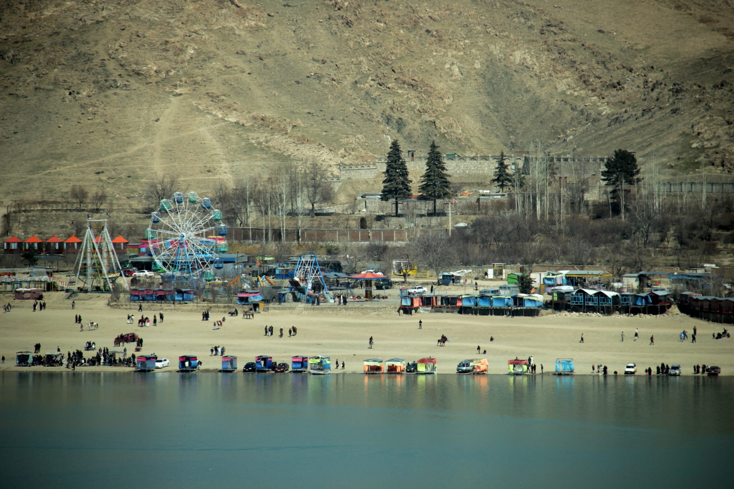 Photograph of colorful ferris wheel and boats alongside a barren mountain. There is lake water in the foreground.