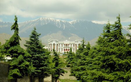 Photograph of green forest trees, in the middle is a historical looking building and a snow-topped mountain range in the background.