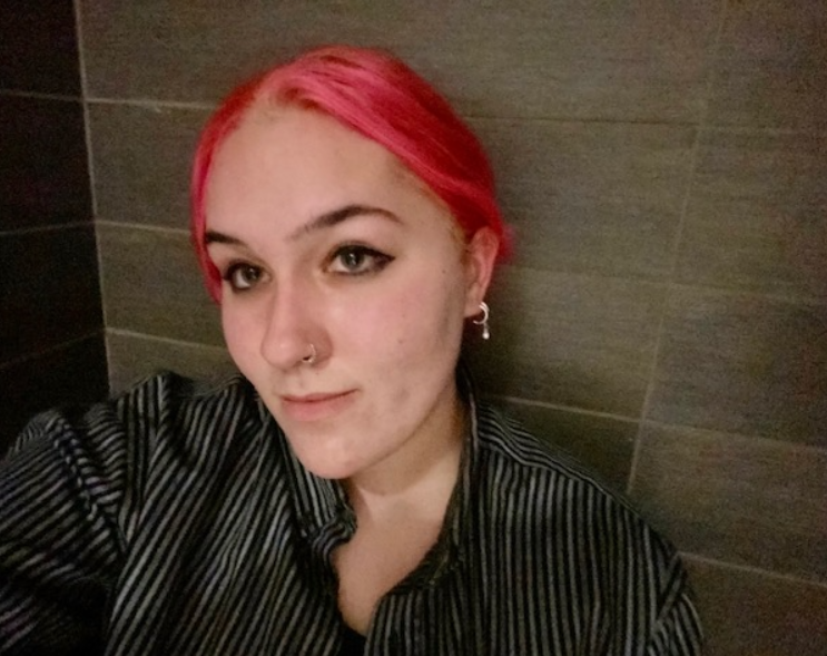 Selfie of a person with pink hair, striped top, and a nose piercing lookup up at camera.